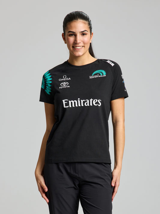 A woman with long dark hair tied back stands smiling at the camera, wearing a SLAM ETNZ DECK WS T-SHIRT in ETNZ BLACK and black pants. The plain, light grey background highlights her Americas Cup pride.