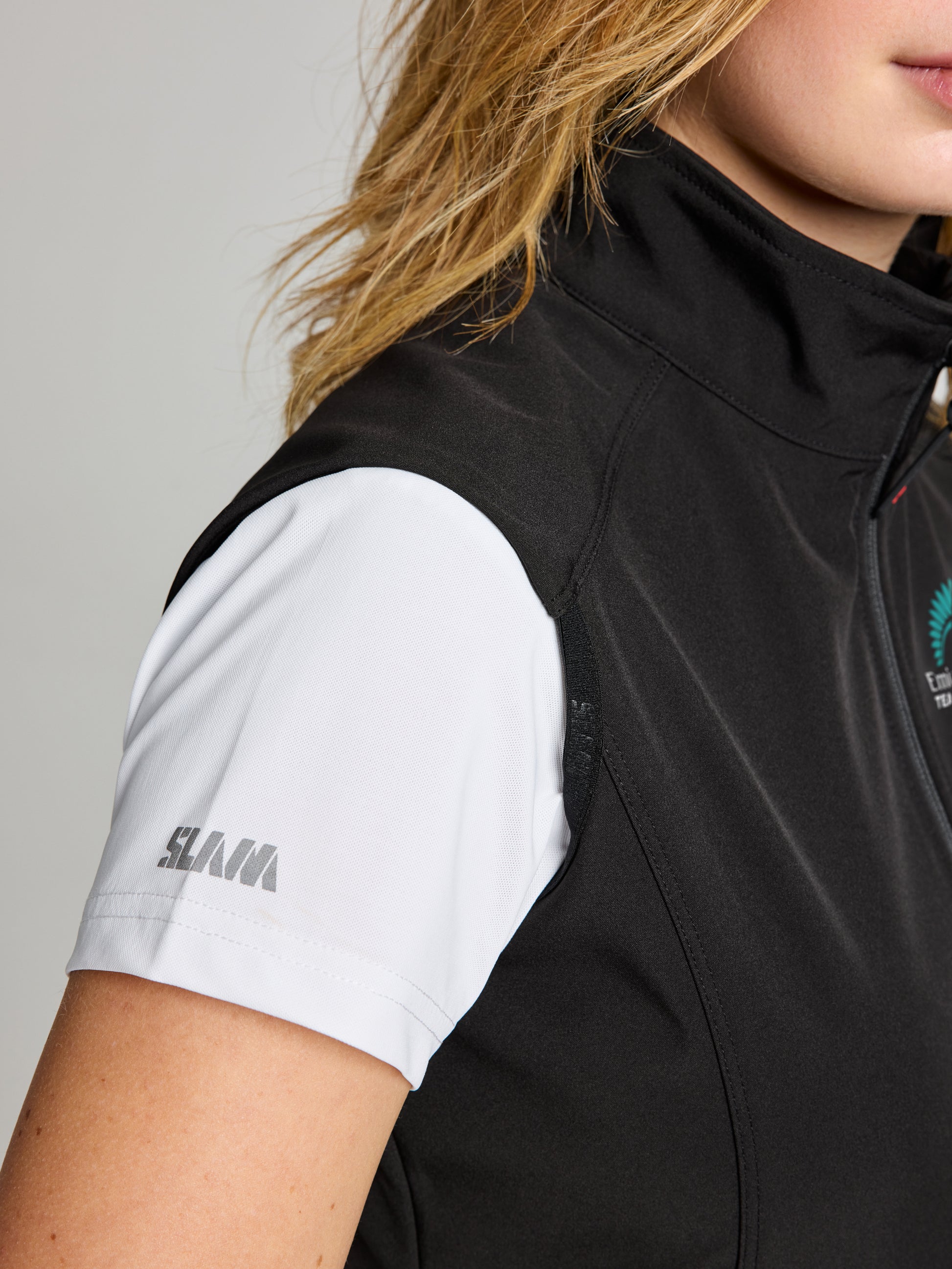 A person is shown from the right side wearing a white SLAM short-sleeved shirt and a black SLAM ETNZ ACT WS SOFTSHELL VES, with only part of their upper body, face, and hair visible, suggesting an Emirates Team New Zealand link.