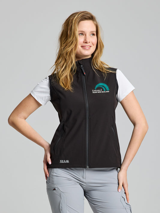 A woman stands against a plain background wearing a SLAM ETNZ ACT WS SOFTSHELL VES in BLACK INK with "Equestrian Team New Zealand" and fern logo, over a white shirt and light gray pants, smiling with one hand on her hip.